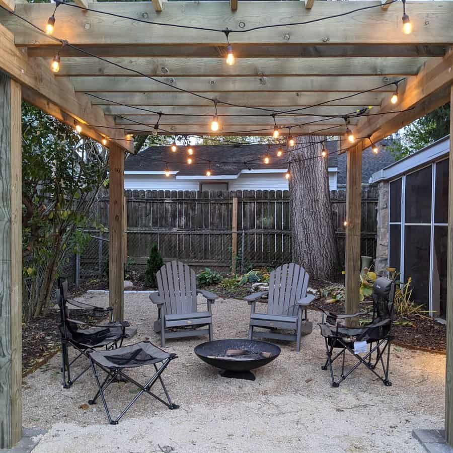 A pergola with string lights covers Adirondack chairs and folding chairs around a fire pit on gravel, with a wooden fence in the background