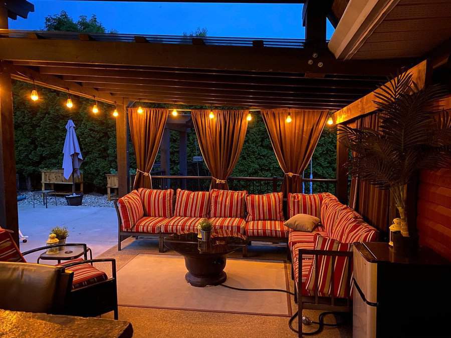 Outdoor patio with string lights, red-striped sofas, and a wooden pergola with curtains. A potted plant sits beside the seating area