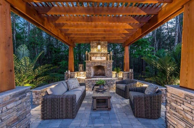 A cozy outdoor seating area with wicker furniture under a wooden pergola, featuring a stone fireplace and lush greenery in the background