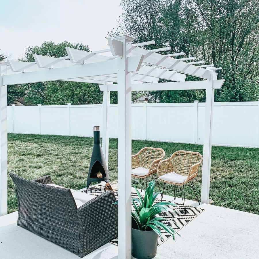 Outdoor patio with a white pergola, wicker chairs, sofa, a chiminea, and potted plant, set on a black and white rug on a concrete floor