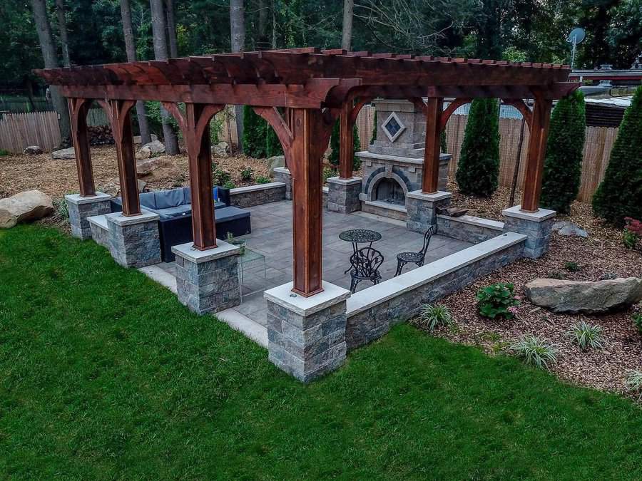 A wooden pergola over a stone patio with fireplace, set in a backyard, features outdoor seating and lush green grass surrounding it