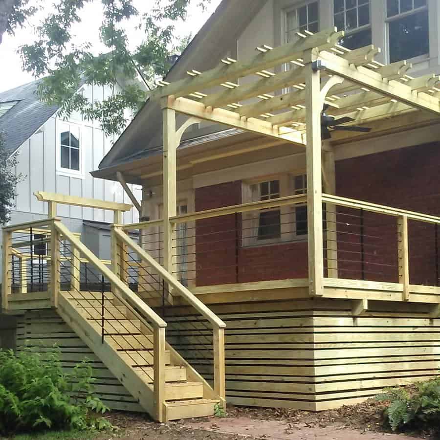 A wooden deck with railing and pergola, attached to a brick house, featuring stairs and surrounded by greenery