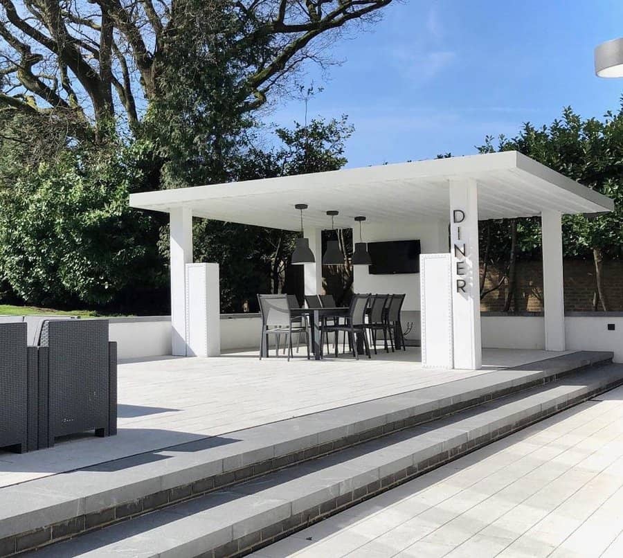 Outdoor dining area with a white pergola labeled "DINER," a table and chairs beneath, surrounded by trees and clear skies