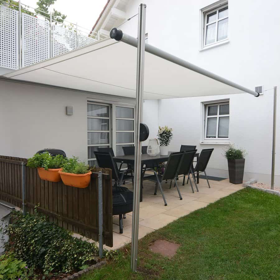 Modern patio with a retractable white shade sail, black dining set, potted plants, and a small garden area