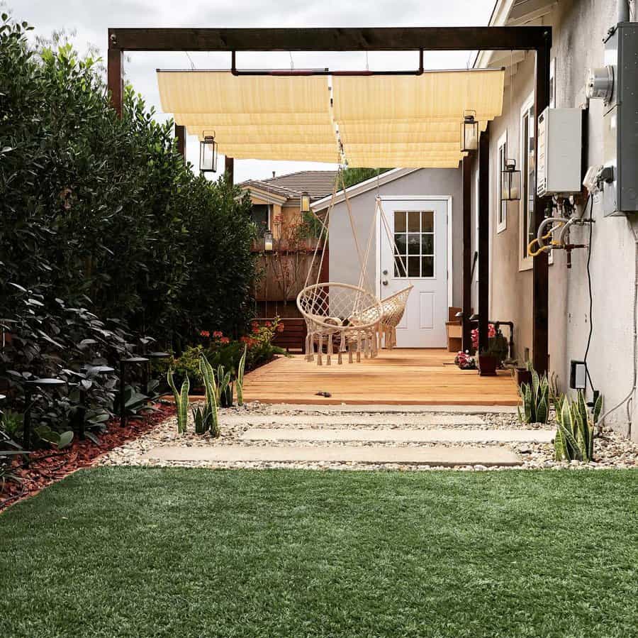 Cozy backyard patio with a retractable pergola shade, hanging macrame chairs, wooden decking, and lush greenery