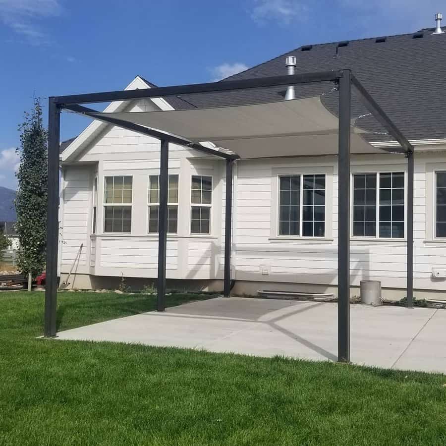 Modern pergola with a white shade sail over a concrete patio, attached to a white house with black trim