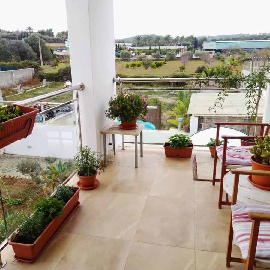 Minimalist balcony garden with potted herbs and flowers, wooden chairs with cushions, and a scenic countryside view through glass railings