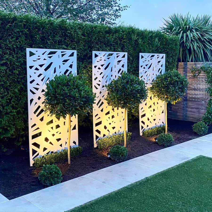 Three decorative white panels with geometric patterns, backlit and flanked by manicured trees in a landscaped garden