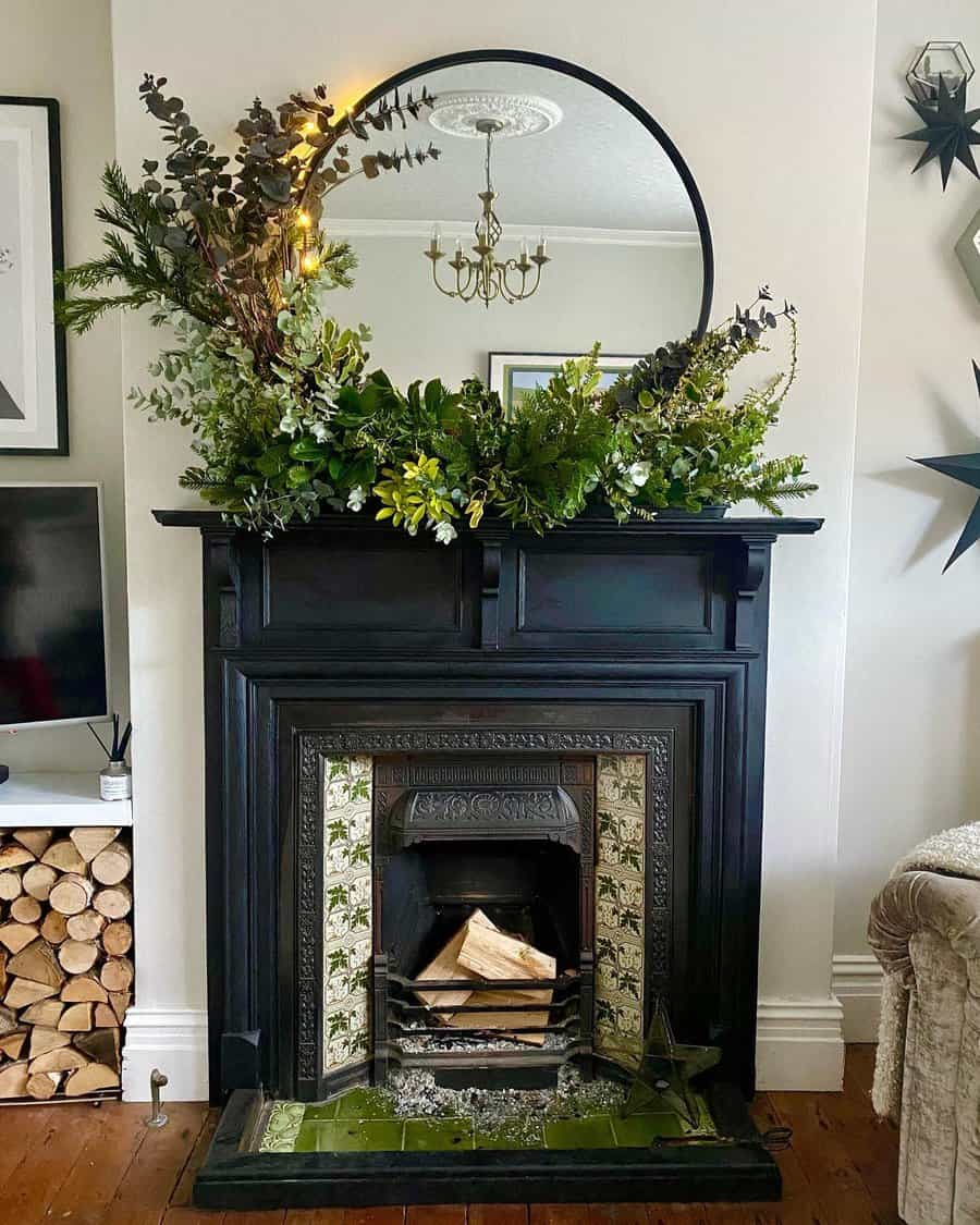 Ornate fireplace with a round mirror, greenery, and logs inside, featuring a chandelier and a pile of firewood visible in the room