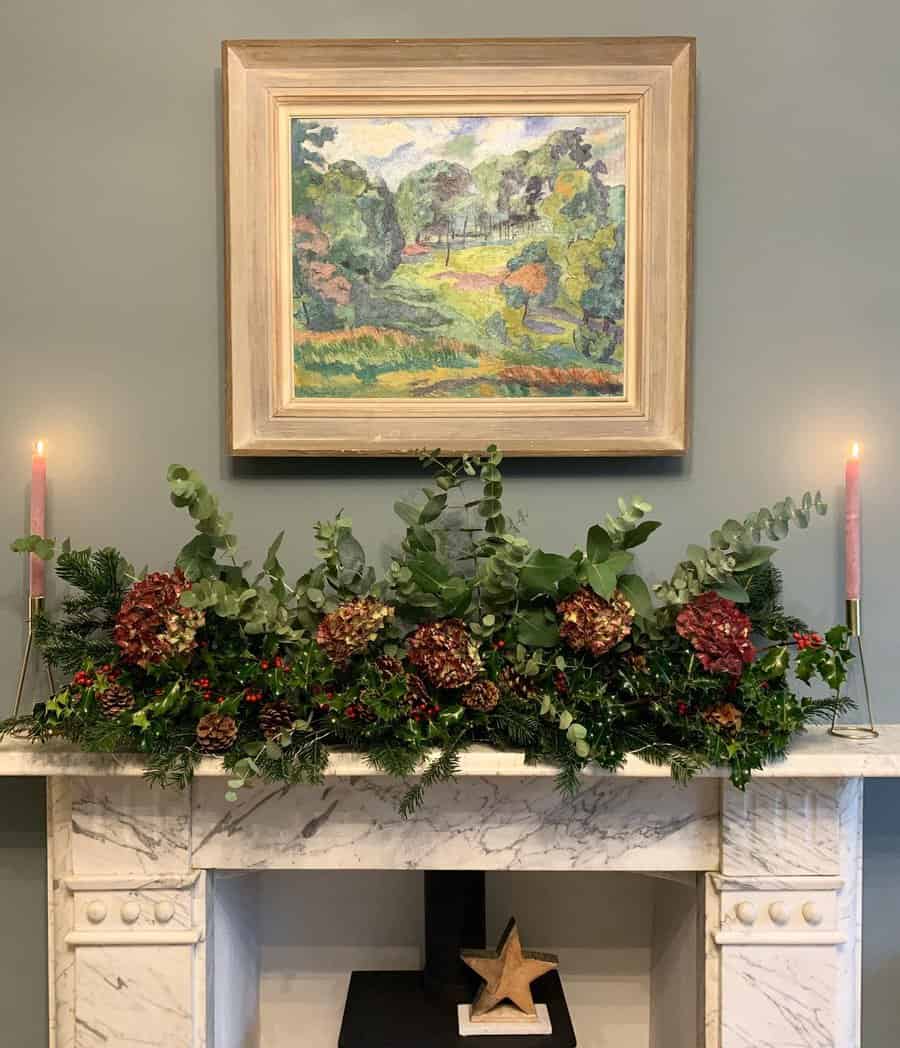 A marble fireplace with greenery, red flowers, candles, and a framed landscape painting above