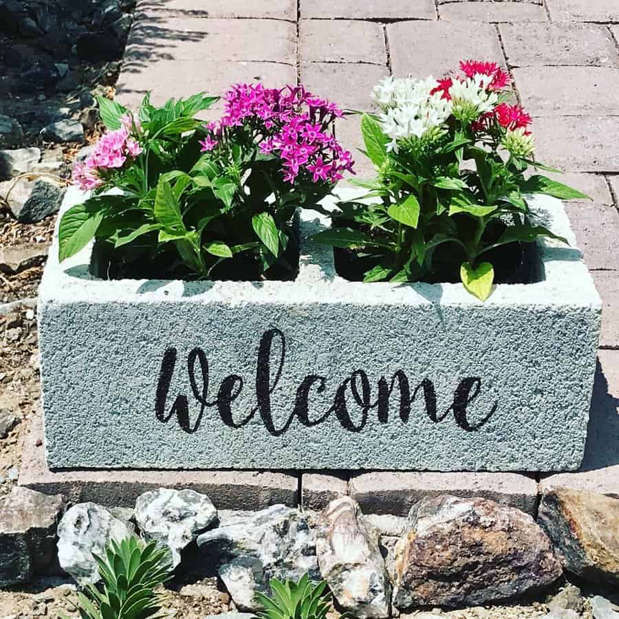 A painted cinder block planter with "Welcome" written on it, filled with colorful flowers, placed on a stone pathway