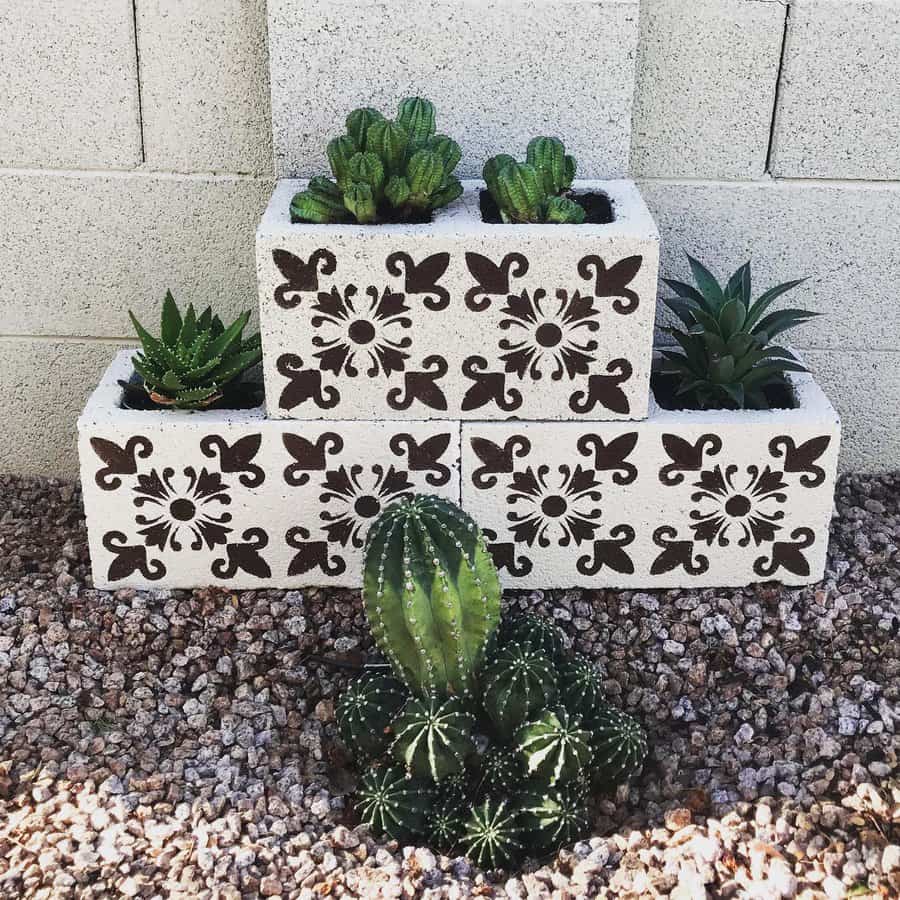 Stacked cinder block planters with intricate black floral designs, filled with succulents and cacti, set against a gravel and brick wall