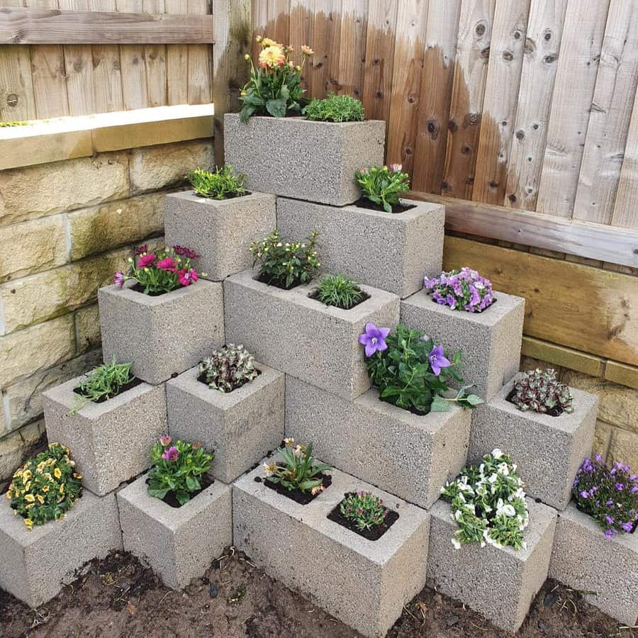 Tiered cinder block planter arranged in a pyramid shape, filled with colorful flowers and greenery, placed in a garden corner against a fence