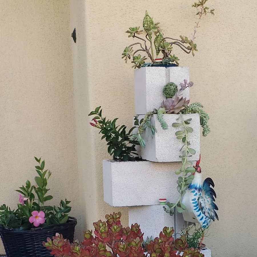 Stacked white cinder block planter with cascading succulents, a ceramic rooster, and potted flowers, set against a beige wall