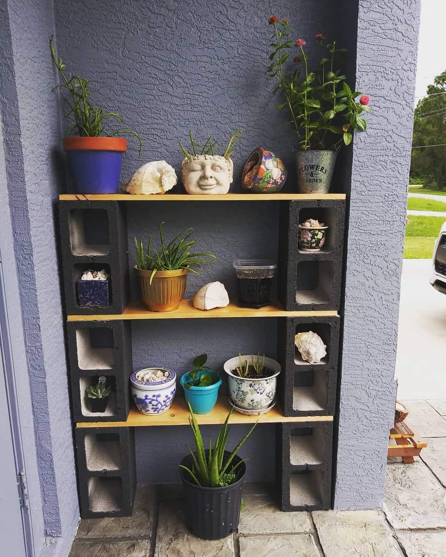 Outdoor shelving with potted plants on cinder blocks against a gray wall, featuring various pots and decorative stones