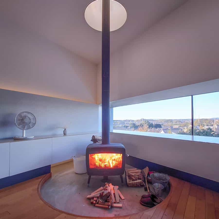 Modern freestanding wood stove on a circular platform, set against panoramic windows with stunning views, creating a warm and minimalist retreat.