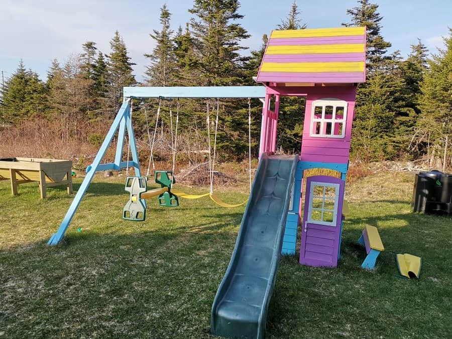 Colorful playground set with slide, swings, and clubhouse on grass, surrounded by trees and picnic table in the background