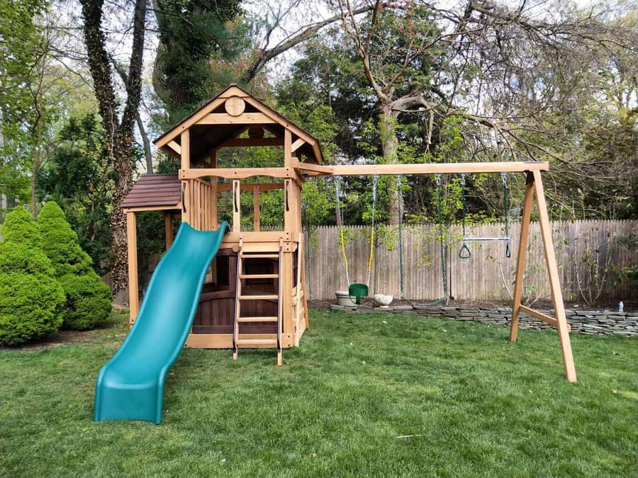 Wooden playset with a green slide, swing set, and climbing ladder in a grassy backyard surrounded by trees and a wooden fence