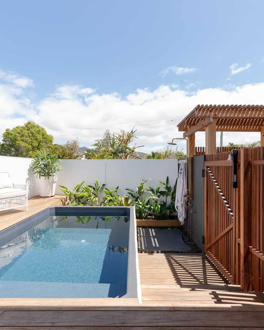 Small rectangular pool with surrounding plants, wooden deck, and privacy fence on a sunny day with blue sky and light clouds