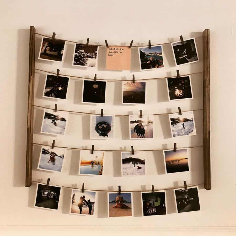 A wooden frame hangs on the wall with four rows of photos clipped to strings, displaying various outdoor scenes and moments