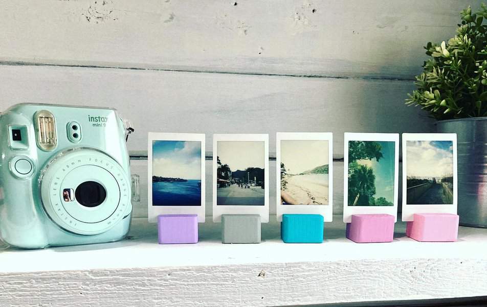 A light blue instant camera and five Polaroid photos on wooden stands on a shelf, with a small plant in the background