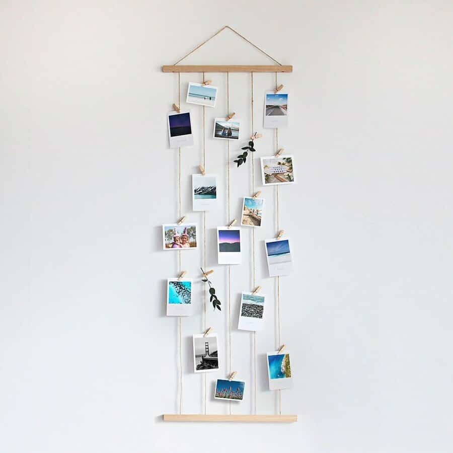 Wall art display with hanging photos clipped on strings between two wooden rods, adorned with small green leaves