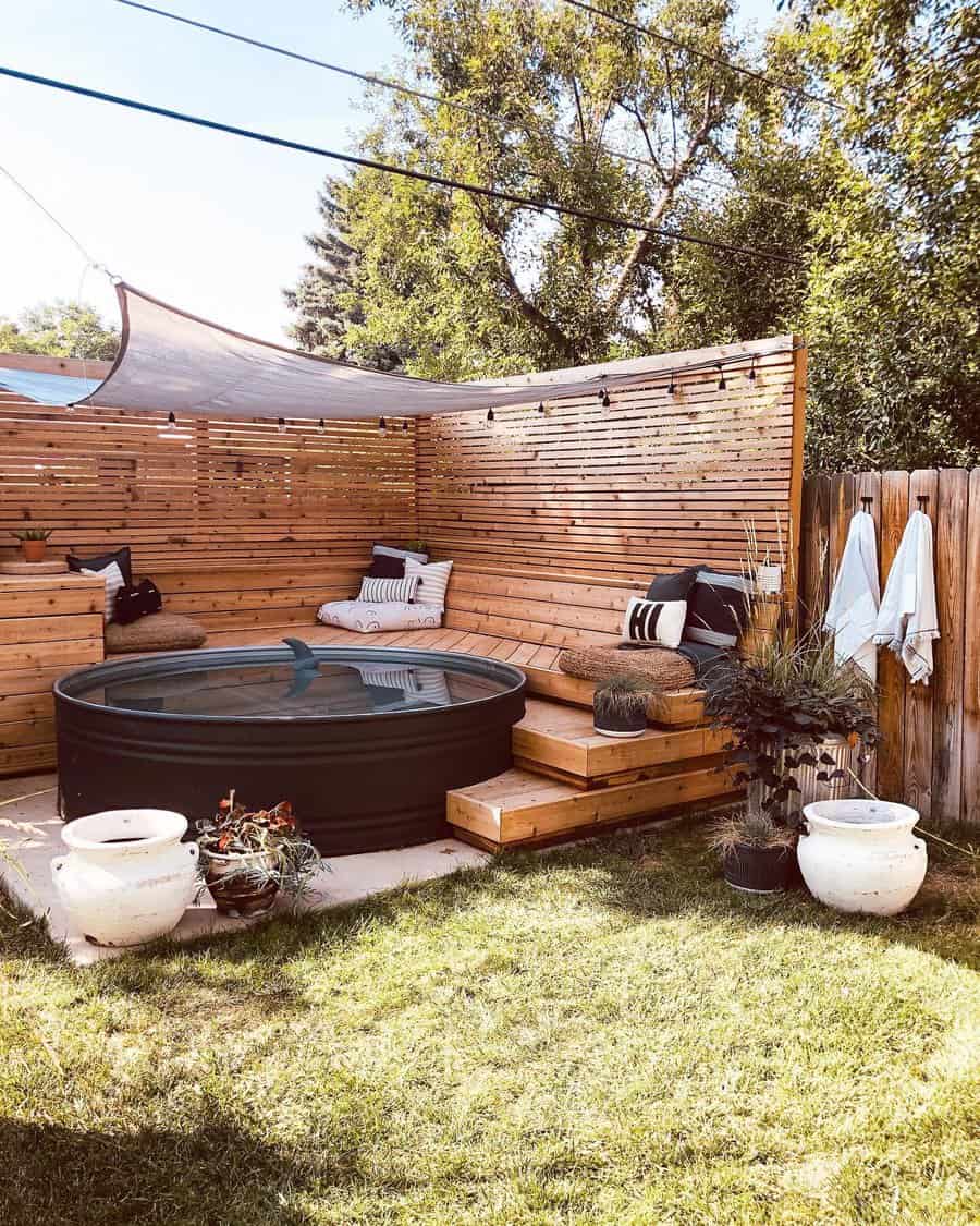 Cozy outdoor deck with a round pool, wooden seating, pillows, and greenery, surrounded by a slatted wooden fence and sunshade