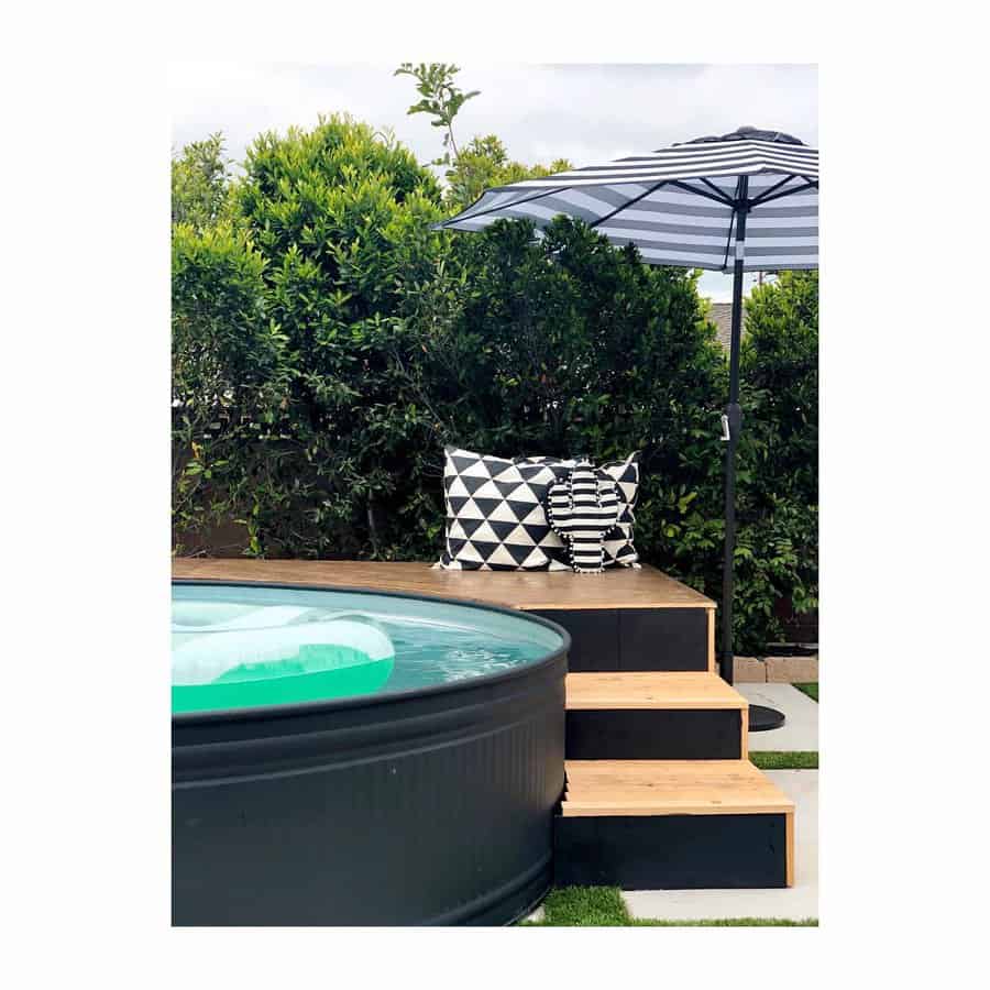 Outdoor stock tank pool area with steps, black and white cushions, and a striped umbrella surrounded by greenery