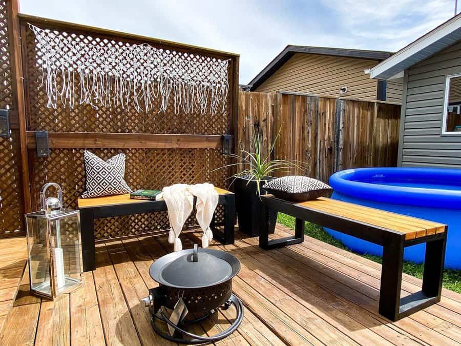 Cozy patio with two wooden benches, a fire pit, decorative pillows, lantern, and a small pool; wooden fence and plant in the background