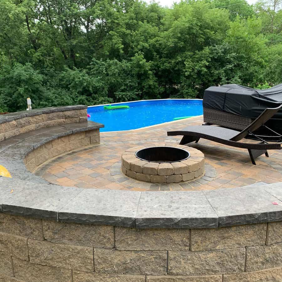 Backyard oasis with a stone paver patio, built-in fire pit, curved seating, and a pool surrounded by lush greenery for ultimate relaxation