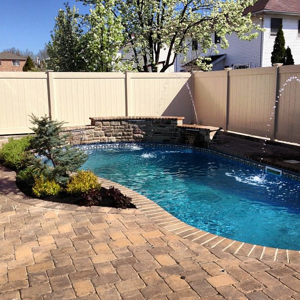 Private backyard pool with warm-toned paver decking, a stone retaining wall, water features, and lush greenery for a tranquil outdoor retreat