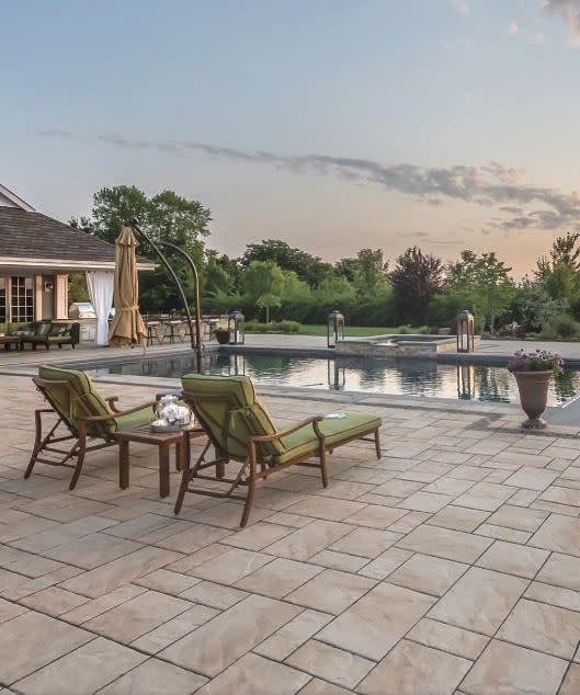 Elegant backyard with a luxurious pool, warm-toned stone pavers, cushioned lounge chairs, and lush greenery for a serene outdoor retreat