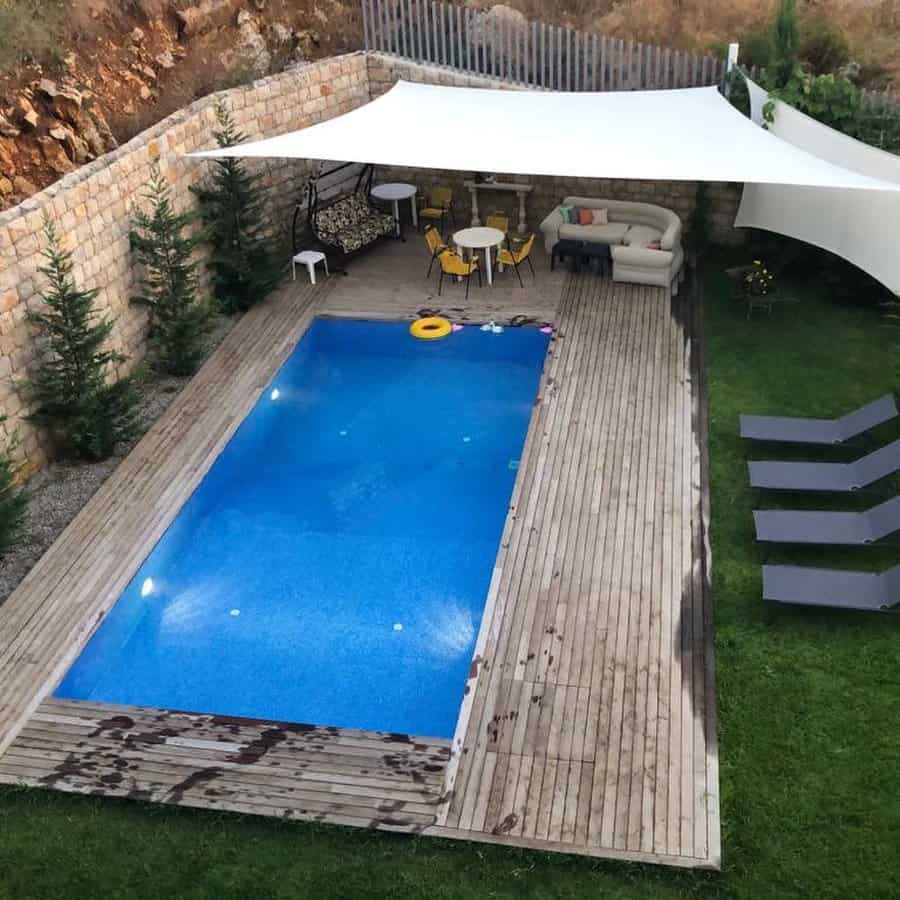 Backyard pool area with a white shade sail, wooden deck, lounge chairs, outdoor seating, and stone wall landscaping