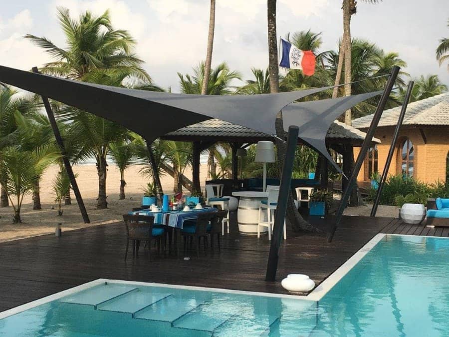 Luxury beachfront pool area with modern black shade sails, wooden deck, stylish dining setup, and tropical palm trees