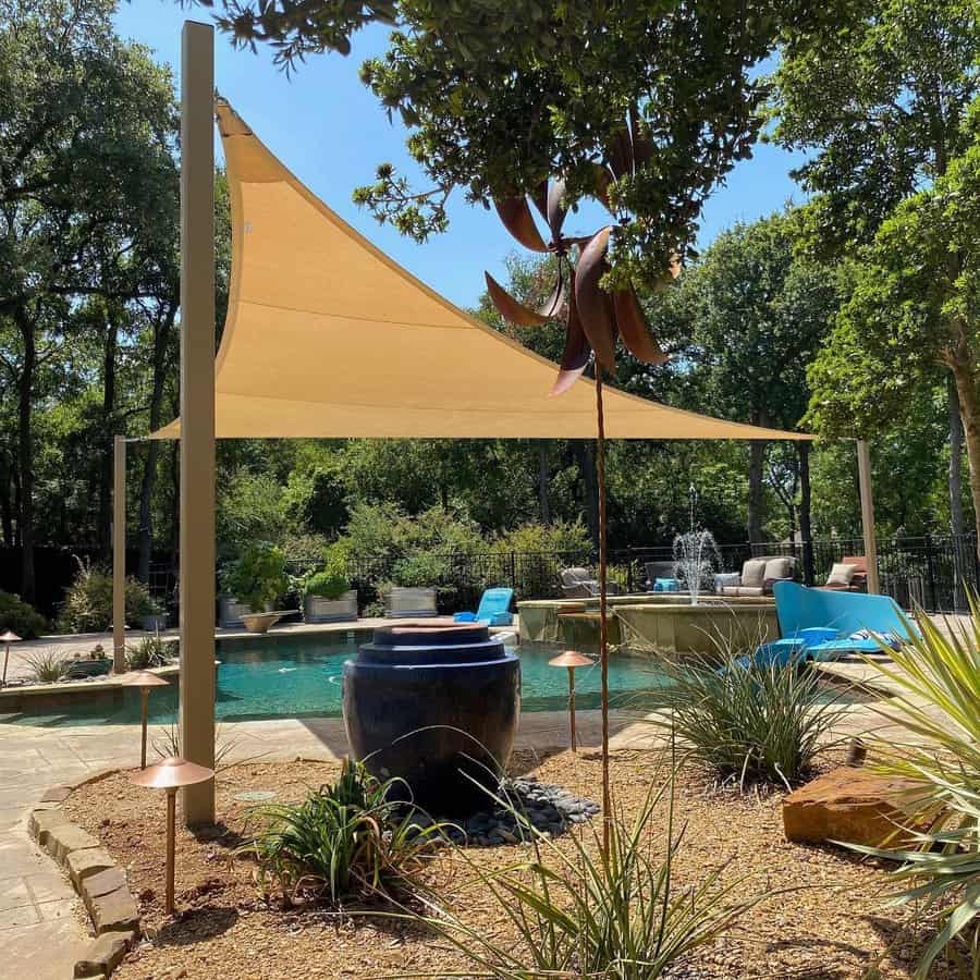 Backyard pool with a tan shade sail, water fountain, lounge chairs, and lush landscaping for a serene outdoor retreat