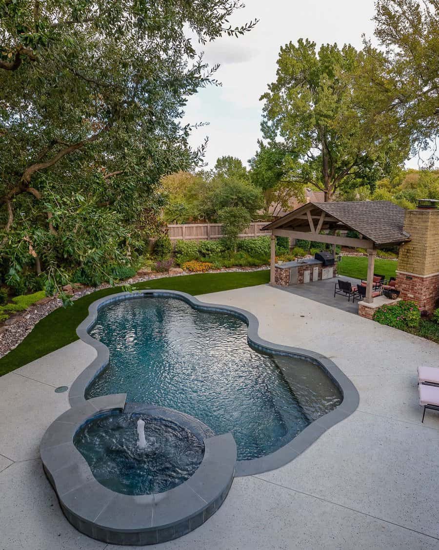 Aerial view of a backyard with a uniquely shaped pool, adjacent hot tub, patio area with a pergola, and surrounding greenery