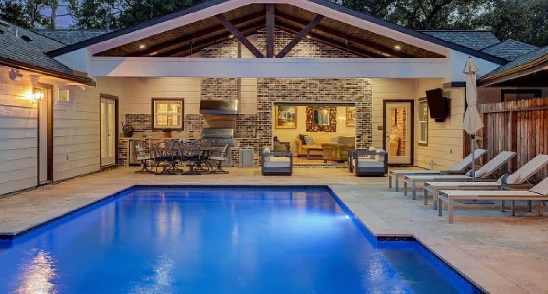 A well-lit backyard pool with a covered patio area, featuring lounge chairs, a dining set, and a cozy seating area