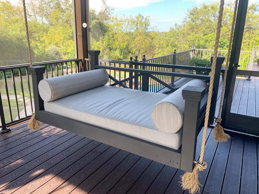 Outdoor porch swing bed with a cushioned seat and bolster pillows, suspended by ropes, with a view of trees and greenery beyond