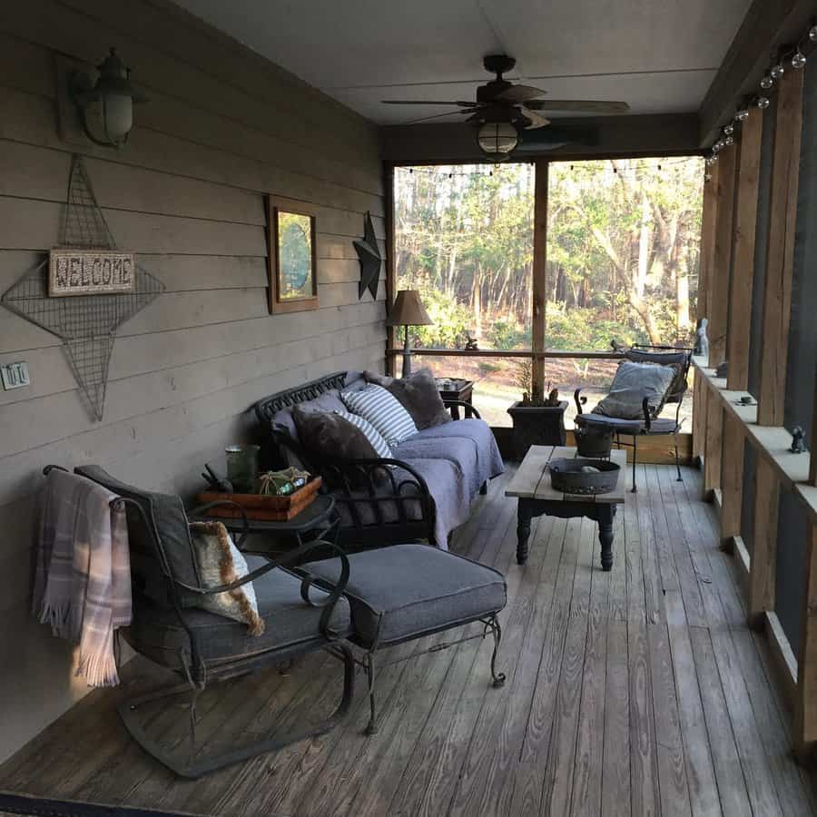 Cozy screened porch with cushioned chairs, tables, and wall decorations; wood flooring and forest view in the background