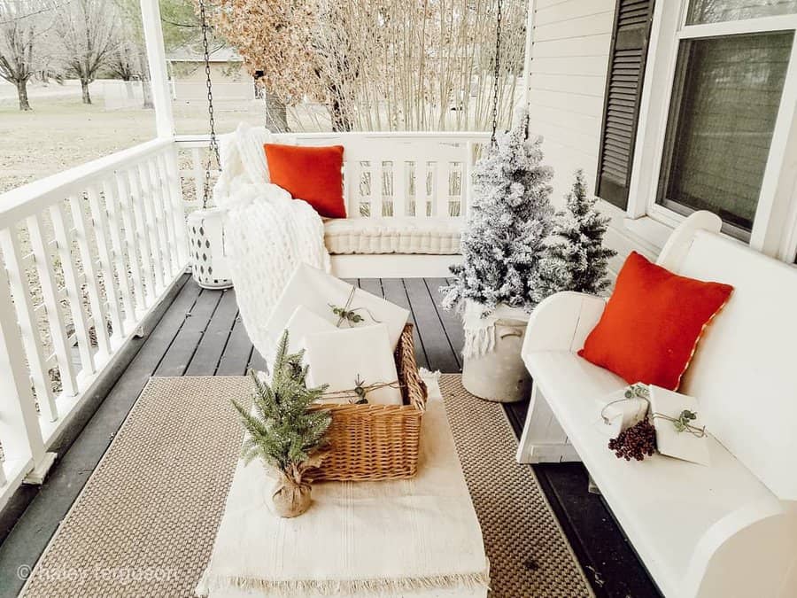 Cozy porch with holiday decor, white furniture, red pillows, and small Christmas trees