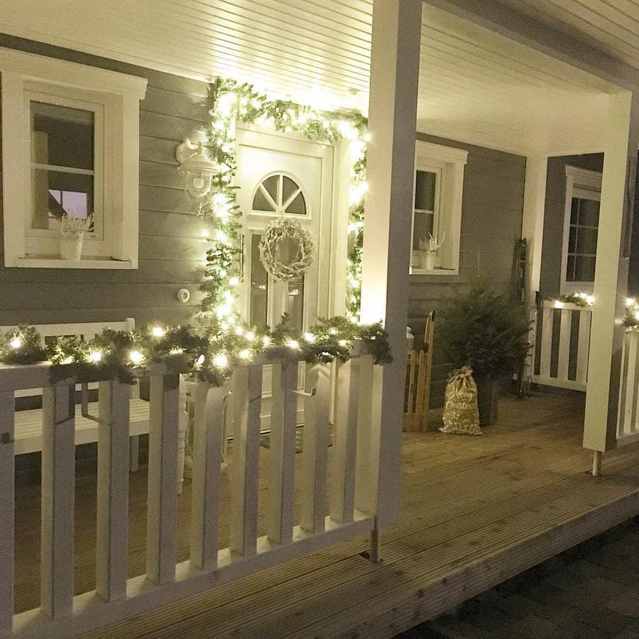 Cozy porch with festive white lights adorning the railing and door, small evergreens, and a decorative sled by the entrance