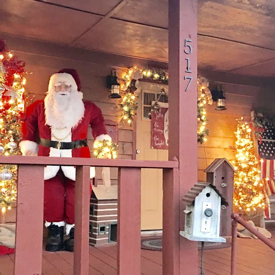 Santa figure stands on a decorated porch with lights, wreaths, and a birdhouse; house number 517 is visible; American flag hangs nearby