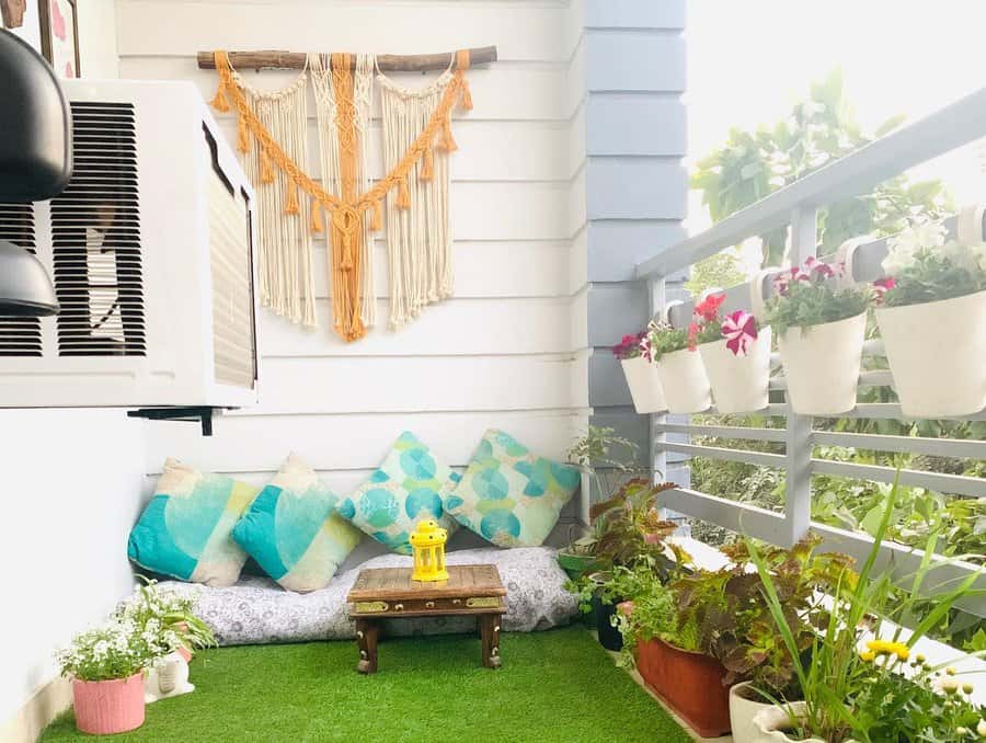 Cozy balcony with green turf, colorful cushions, a small table with a yellow lantern, potted plants, and wall macramé decor