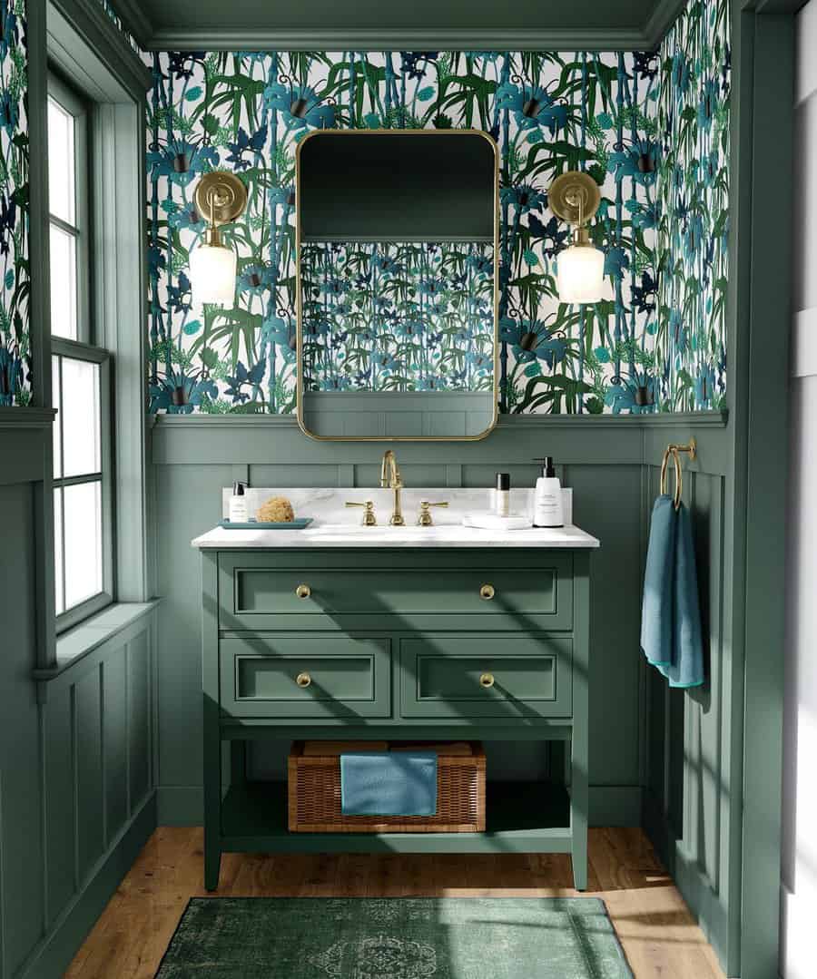 A bathroom with floral wallpaper, green vanity with brass fixtures, oval mirror, dual wall sconces, and a green towel and rug