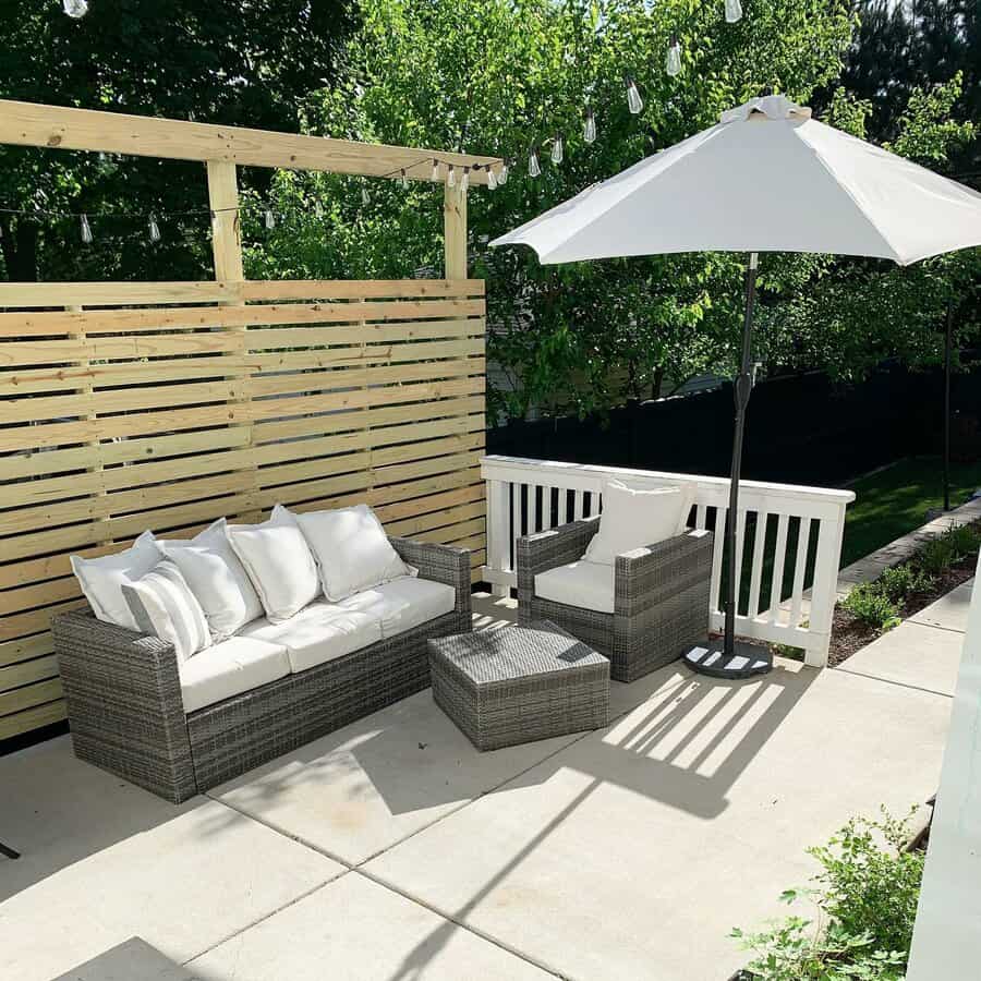 Patio with wicker furniture, white cushions, a square table, umbrella, and string lights set against a wooden privacy screen