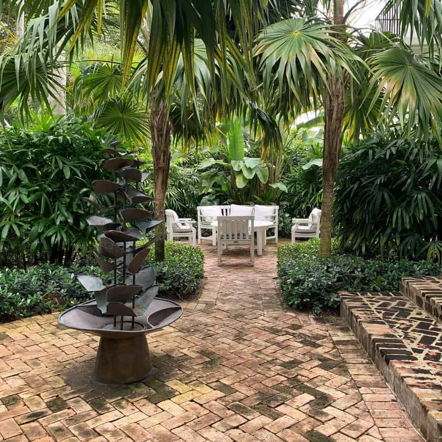 Secluded Florida garden with tropical palms, lush greenery, a brick pathway, and a modern water fountain leading to a cozy outdoor dining space