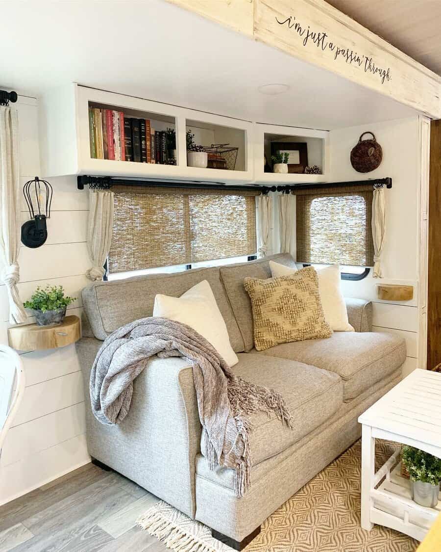 Cozy RV interior with a grey sofa, pillows, blanket, mounted shelves with books, wicker blinds, and plants