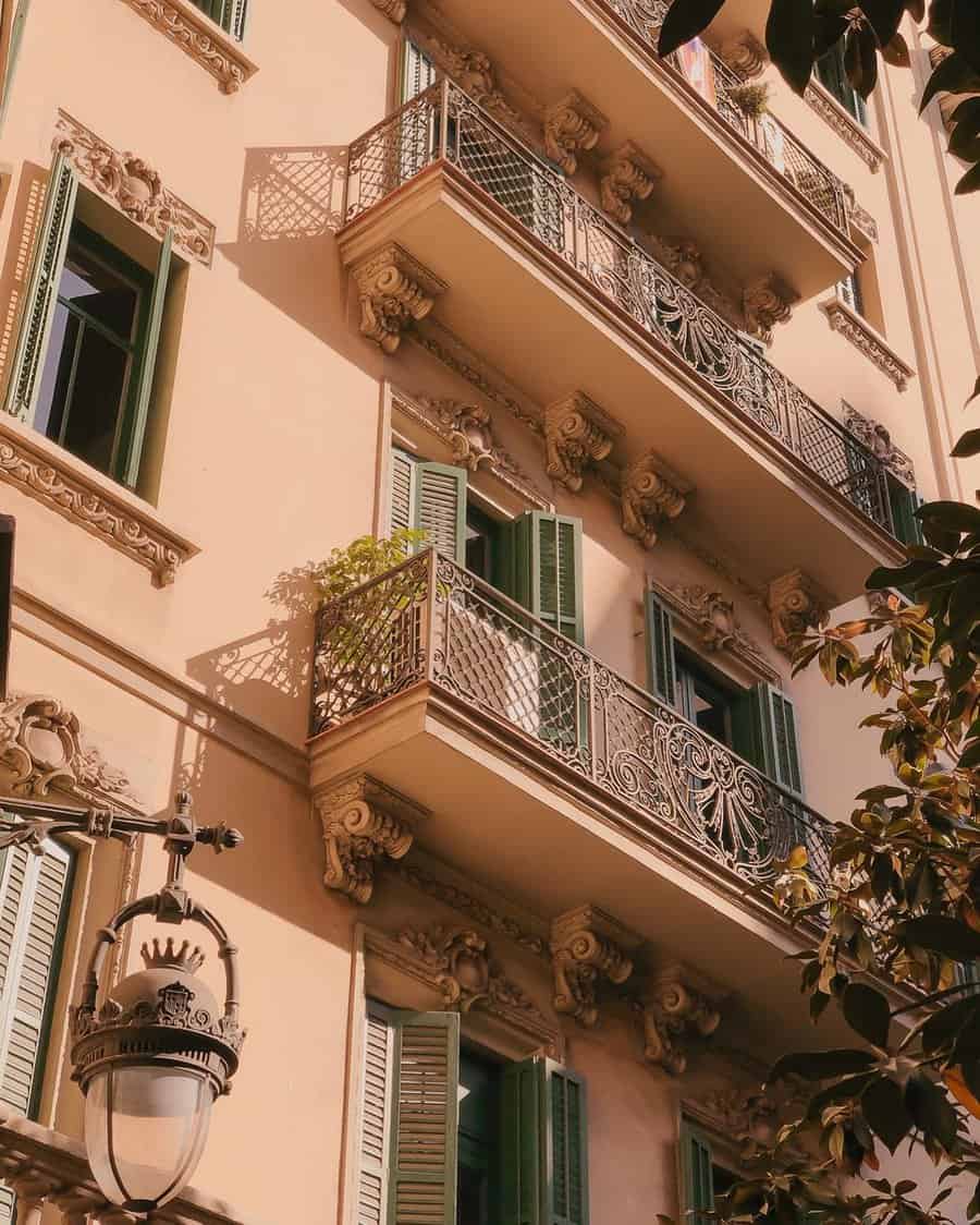 Ornate building facade with green shutters and decorative iron balconies, tall lantern in foreground, sunlit with leafy shadows