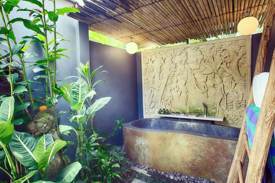 Outdoor bathroom with a stone bathtub, bamboo roof, lush plants, and a carved stone wall