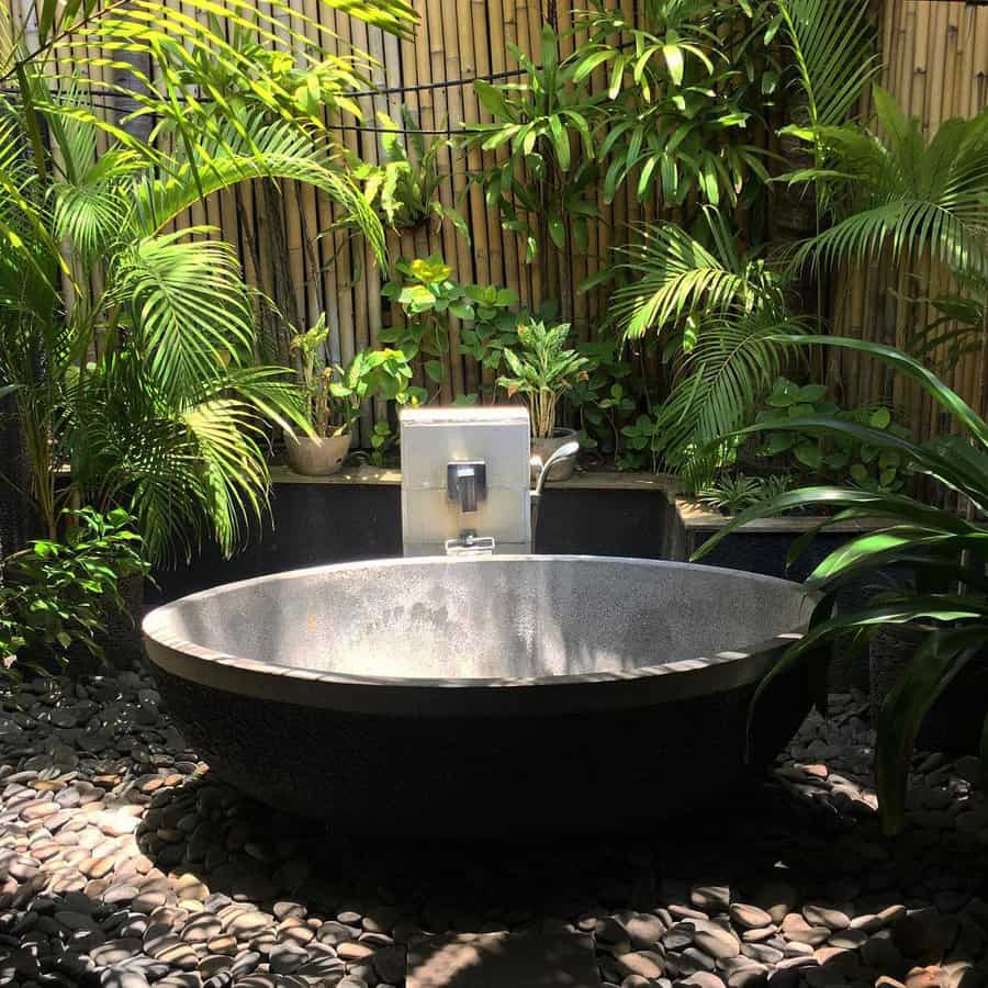 Outdoor stone bathtub surrounded by lush green plants and bamboo fence, with rocks covering the ground to create a serene, natural setting